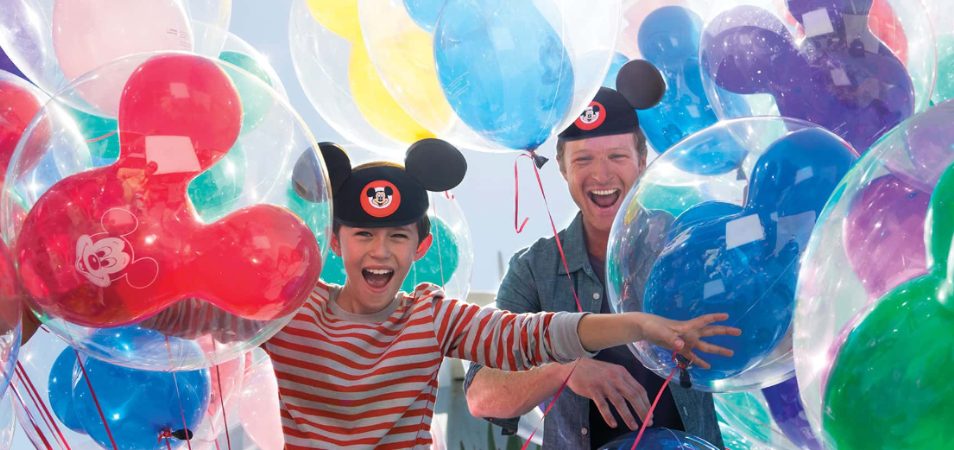 A kid and his dad wearing Mickey Mouse hats surrounded by colorful balloons at Disneyland.