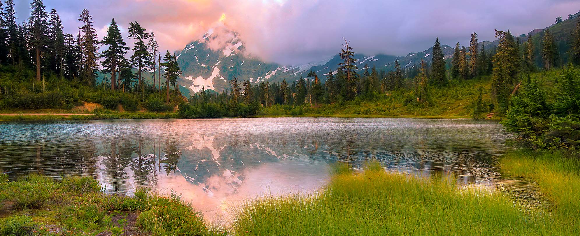A lake, trees, and Mount Shuksan covered in clouds at sunset.