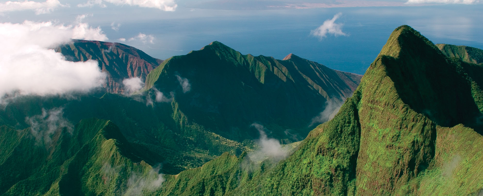 A bird's-eye-view of lush volcanic mountains, the ocean in the distance. 