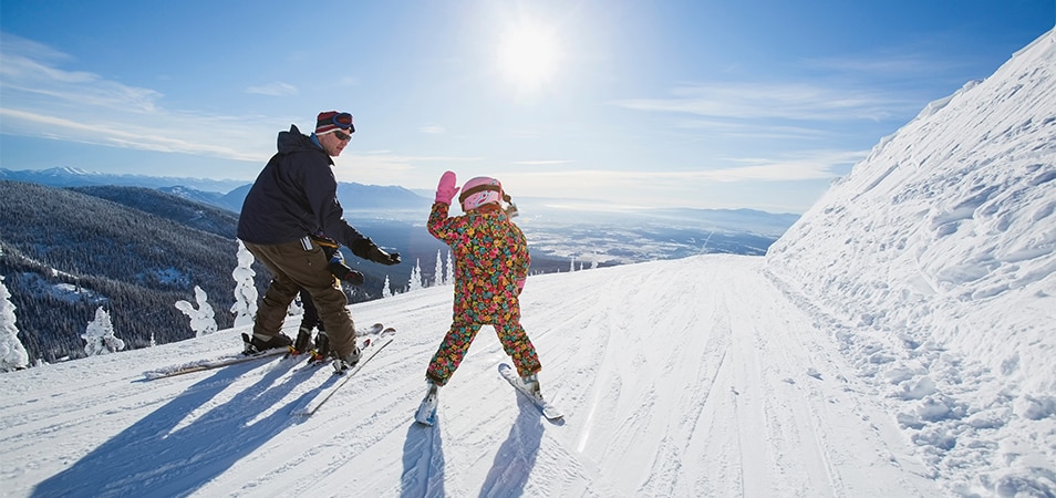 A female child is high-fiving her dad, as they ski down a mountain.