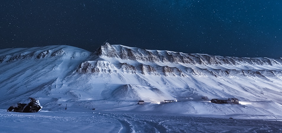 A snowmobile sits in front of a snowy mountain under a starry night sky.