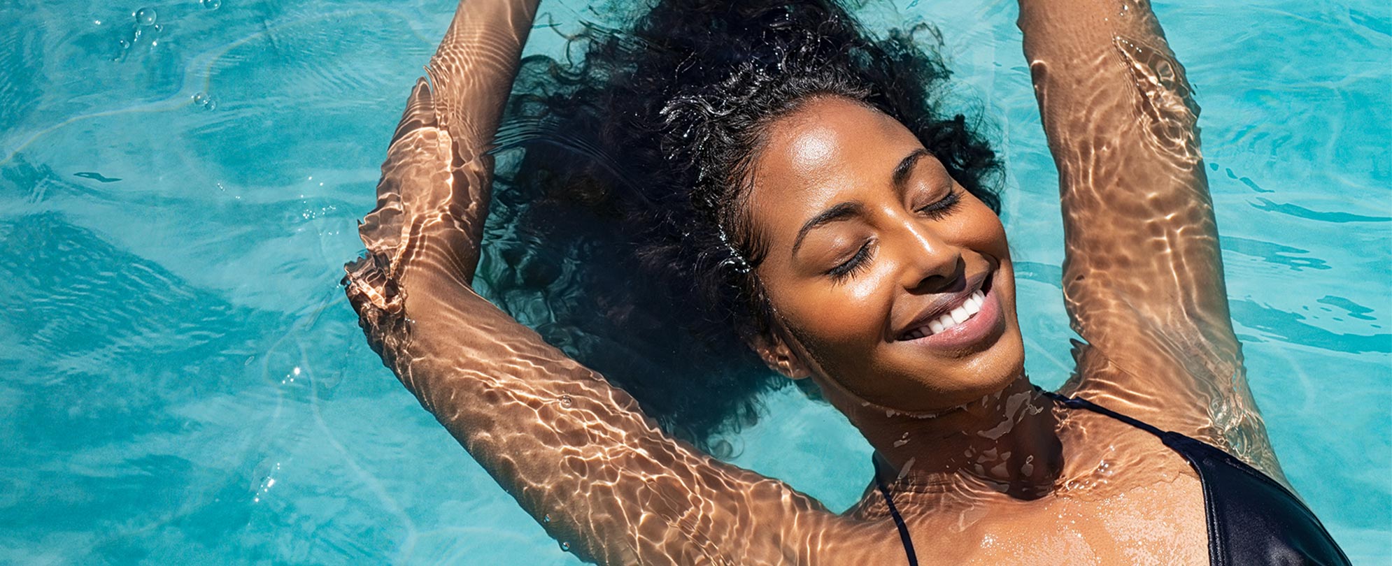 A smiling, African American woman relaxes with her eyes closed floating in a pool.