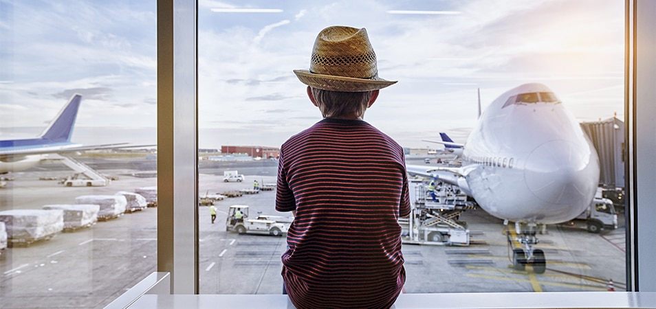 A young boy wearing a hat, looking out of an airport window at a nearby plane. 