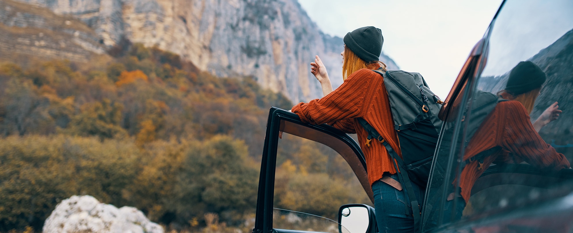 A woman leaning out of her car, pointing at a nearby mountain with trees cascading down.