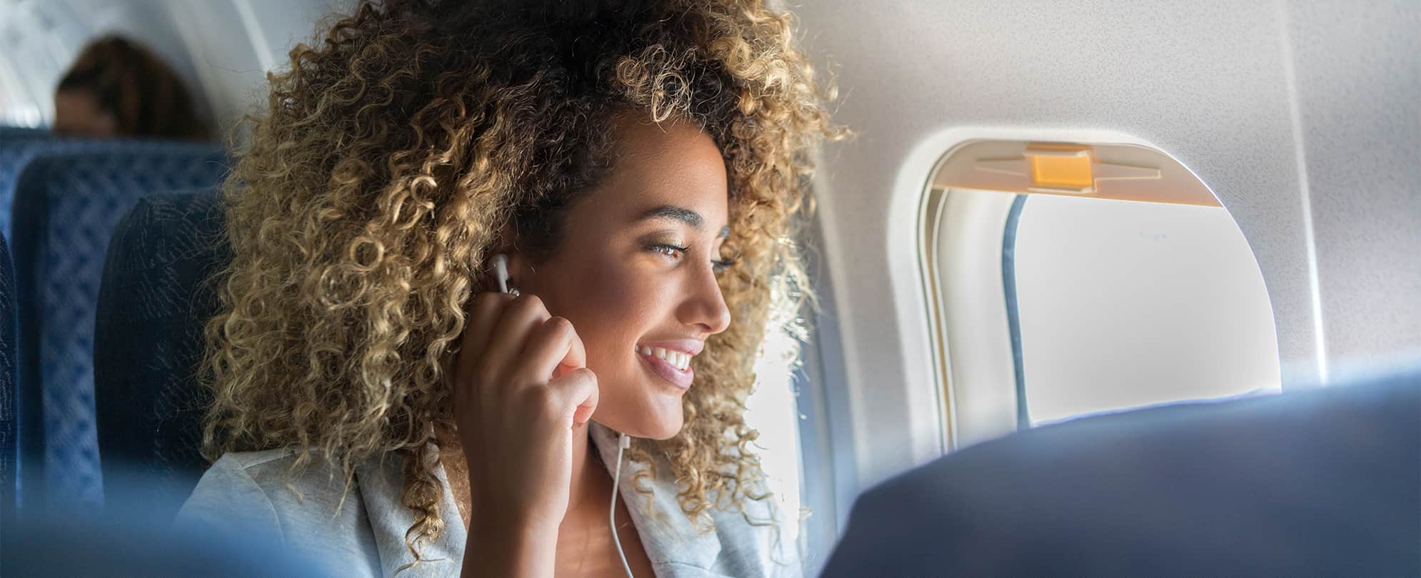 A woman with headphones in her ears, looking outside a plane window.
