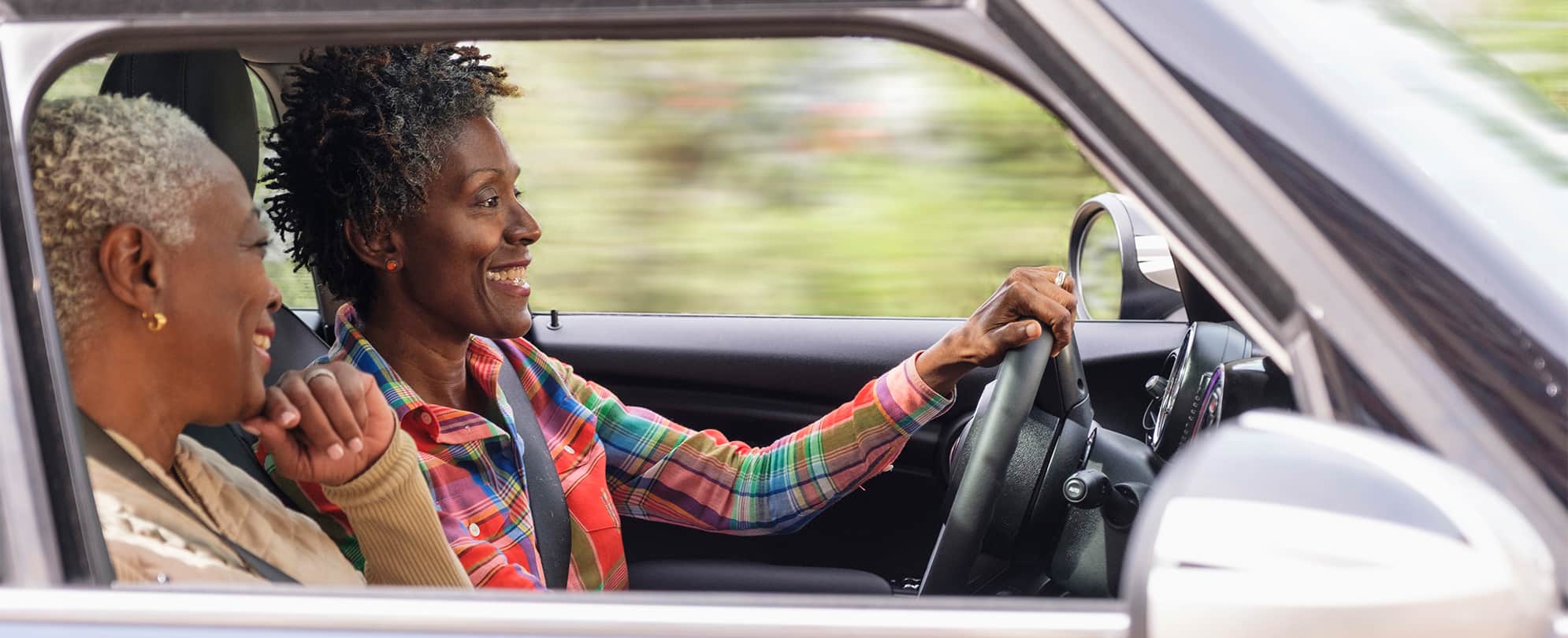 Two smiling women are in a car, one is driving, and the other is in the passenger's seat.