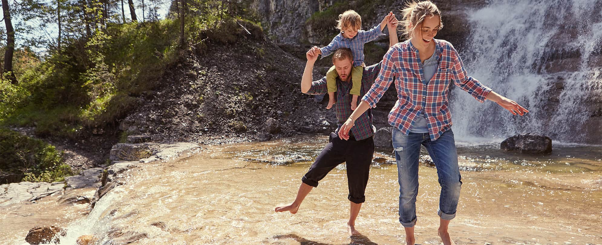 A mother leading her husband and daughter who is on her father's shoulders, across a river barefoot. 