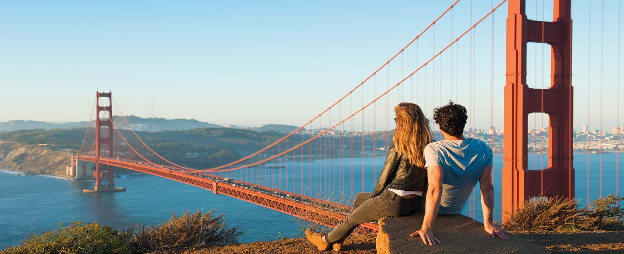 A man and woman sit on a rock up on a hill, staring out over the Golden Gate Bridge in San Francisco, California.