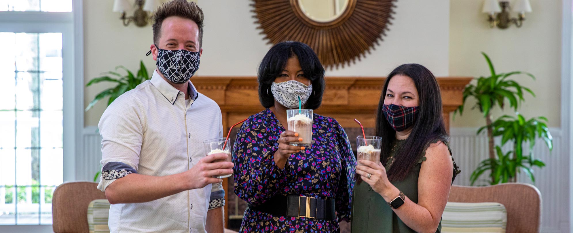 Three friends wearing masks and holding up coffee milkshake drinks with straws.