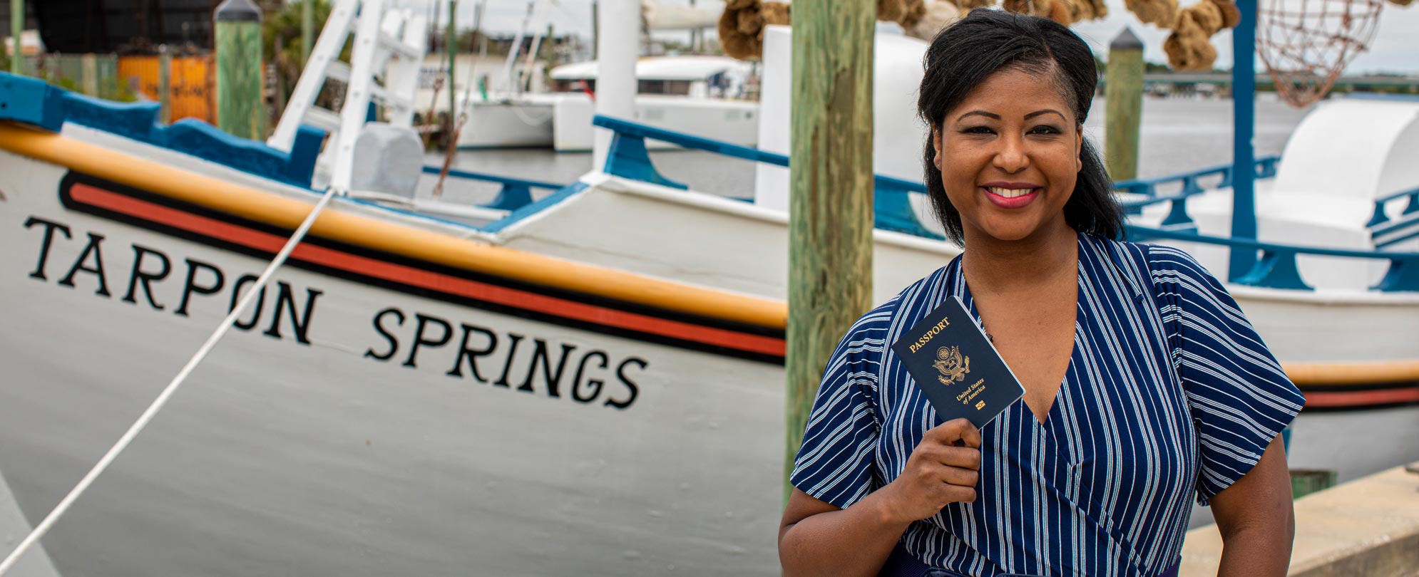 A woman smiling and holding up a passport in front of a Tarpon Springs boat.