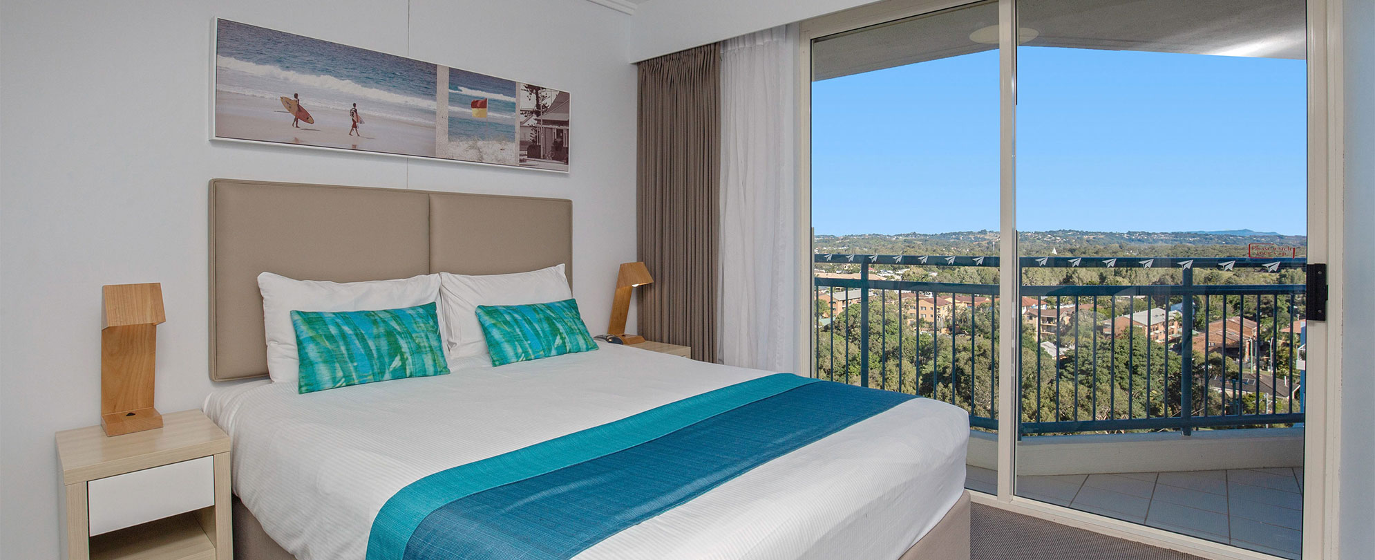 A bedroom with a king bed, 2 nightstands, and a sliding glass door to a balcony in a Club Wyndham Kirra Beach standard suite.