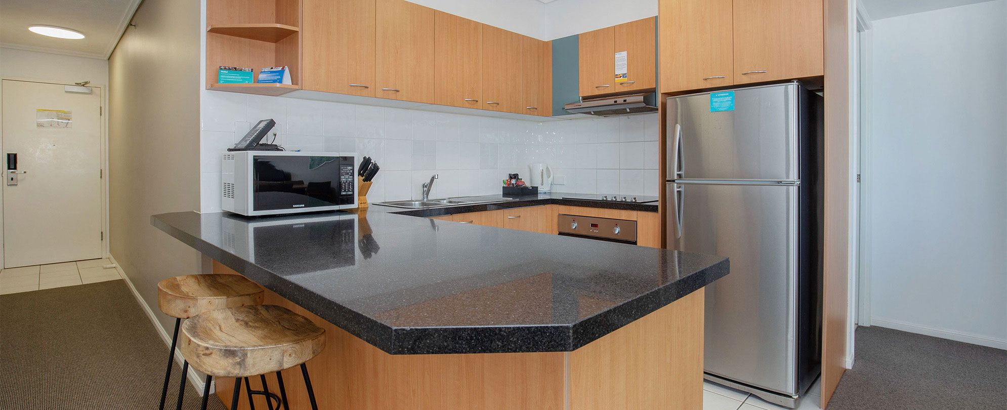 The kitchen of a Club Wyndham Kirra Beach standard suite with a fridge, microwave, stove, and countertop bar with 2 stools.