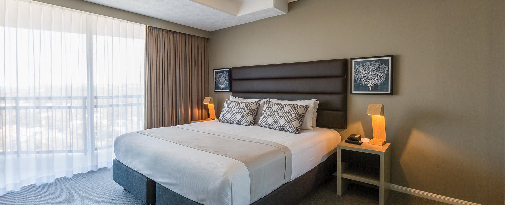 A bedroom with a king bed, 2 nightstands, and a sliding glass door with curtains in a Club Wyndham Kirra beach standard suite.