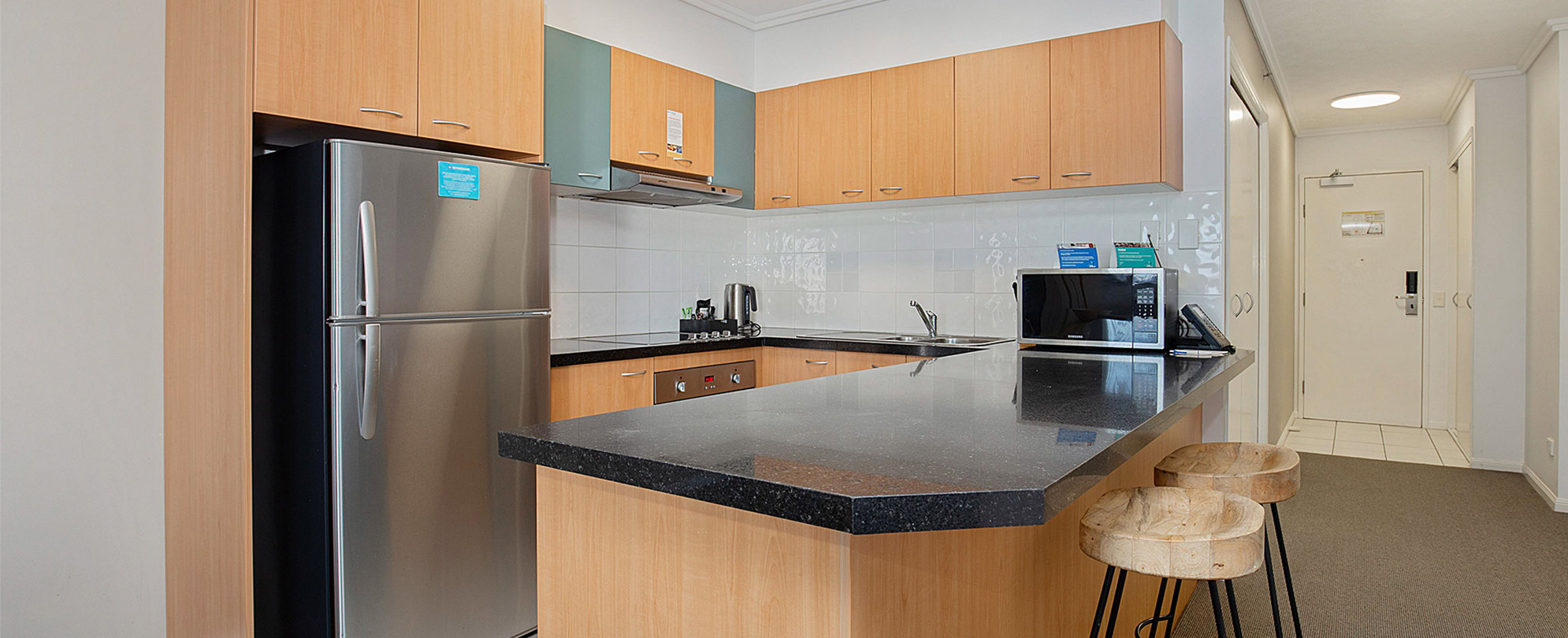 The kitchen of a Club Wyndham Kirra Beach standard suite with a full fridge, microwave, and countertop bar with 2 stools.
