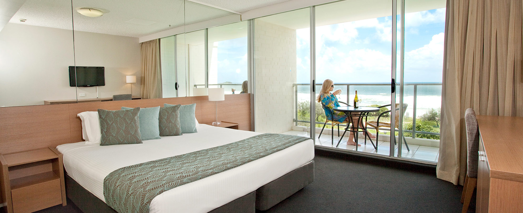 A bedroom with a king bed, mirrored wall, and a sliding glass door to a balcony in a Club Wyndham Marcoola Beach suite. 