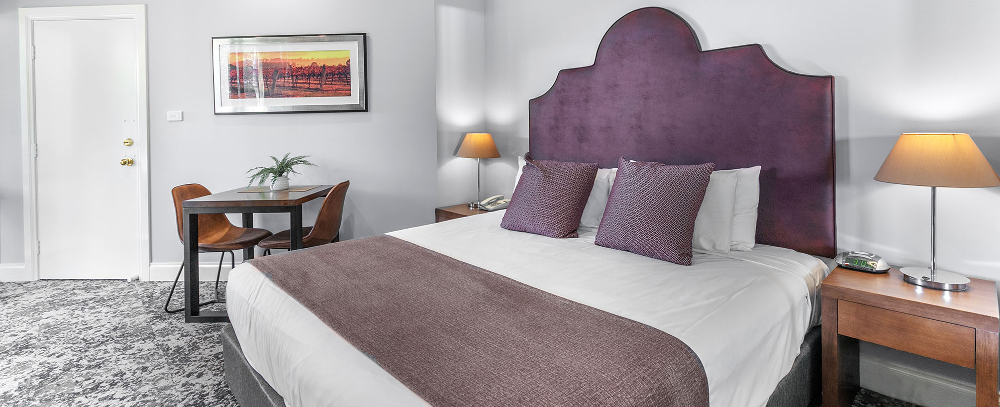 A bed with a headboard, two side tables with lamps, and a small dining table Club Wyndham Pokolbin Hill studio suite.