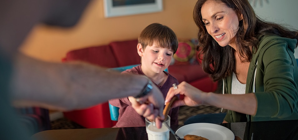 A father is placing a fresh-baked chocolate chip cookie on a plate while a mother and her son dunk their cookies in a glass of milk. 