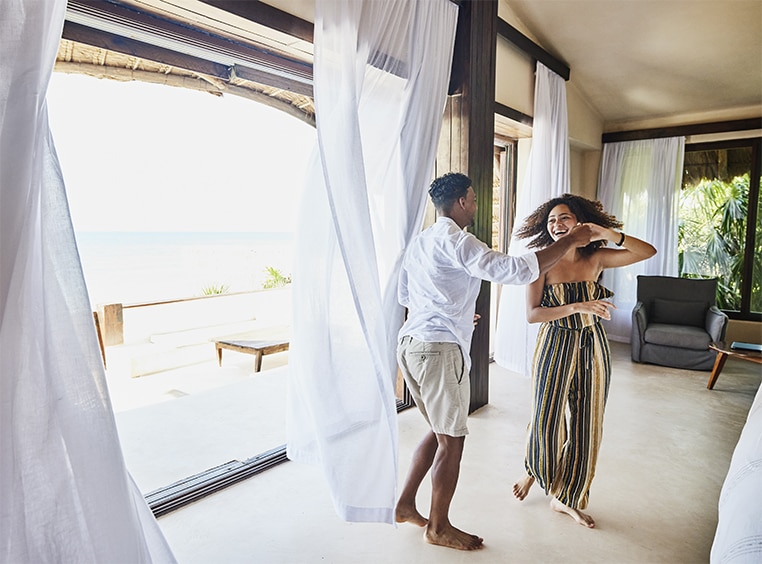 A couple dancing in their oceanfront WorldMark resort suite in front of open windows and doors with long curtains.