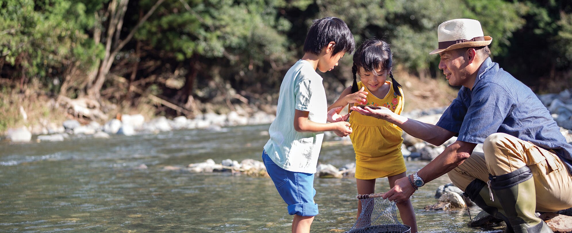 A father and his two kids standing in shallow water, looking at something the father found in the river.