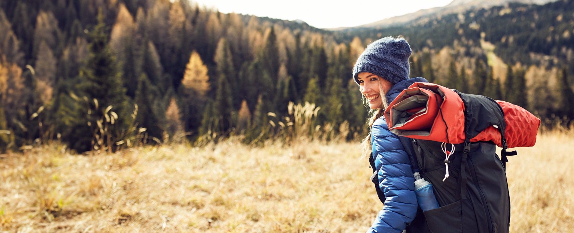 A woman wearing a wool hat and puffer jacket smiles as she looks back while on a sunny winter hike.