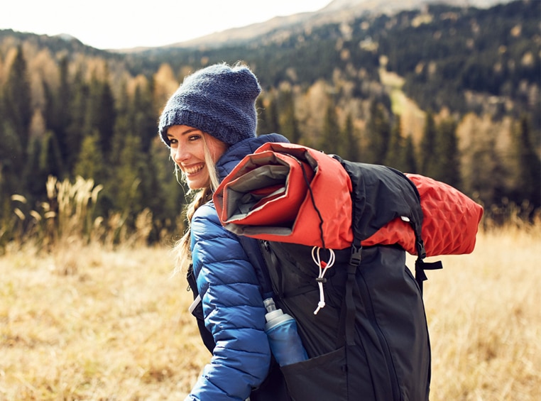 A woman wearing a wool hat and puffer jacket smiles as she looks back while on a sunny winter hike.