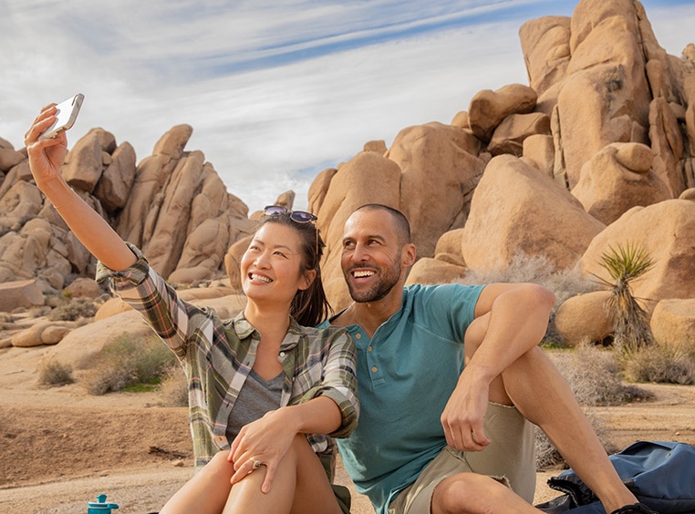 A man and a woman sitting on a rocky path, the woman holds her arm up to take a selfie.