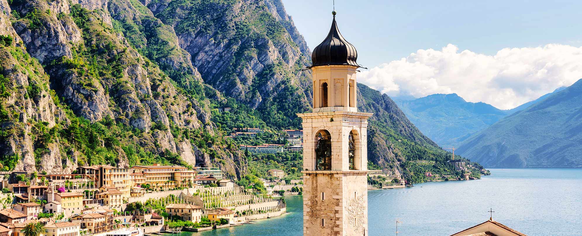 A bell tower in Limone sul Garda, a town in Lombard, Italy, with mountains, buildings, and Lake Garda in the background.