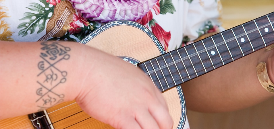 A man with wrist tattoo wearing a tropical print shirt and a lei strums a ukulele.