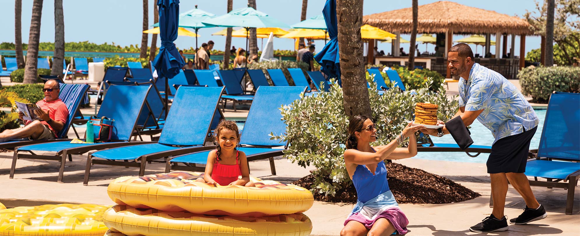A server hands a mom a plate of food while she sits on the edge of a resort pool, next to her daughter on pool floats.