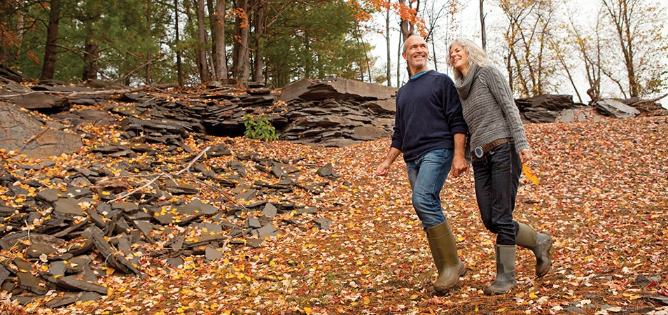 An older couple walks holding hands on a rocky mountain trail covered in fall leaves.