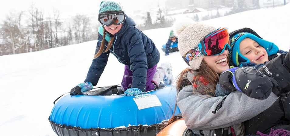 A mom and her young kids wearing winter gear smile while snow tubing.