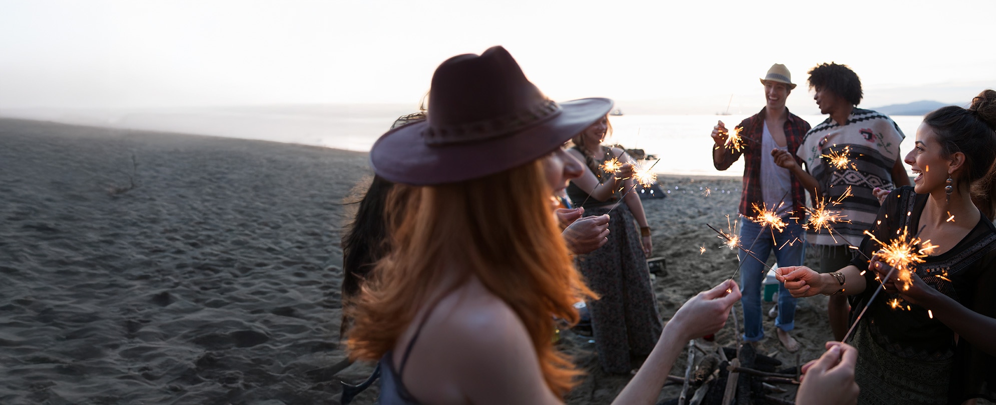 A group of friends gathers on a beach at sunset, joyfully holding sparklers and sharing a festive moment together.