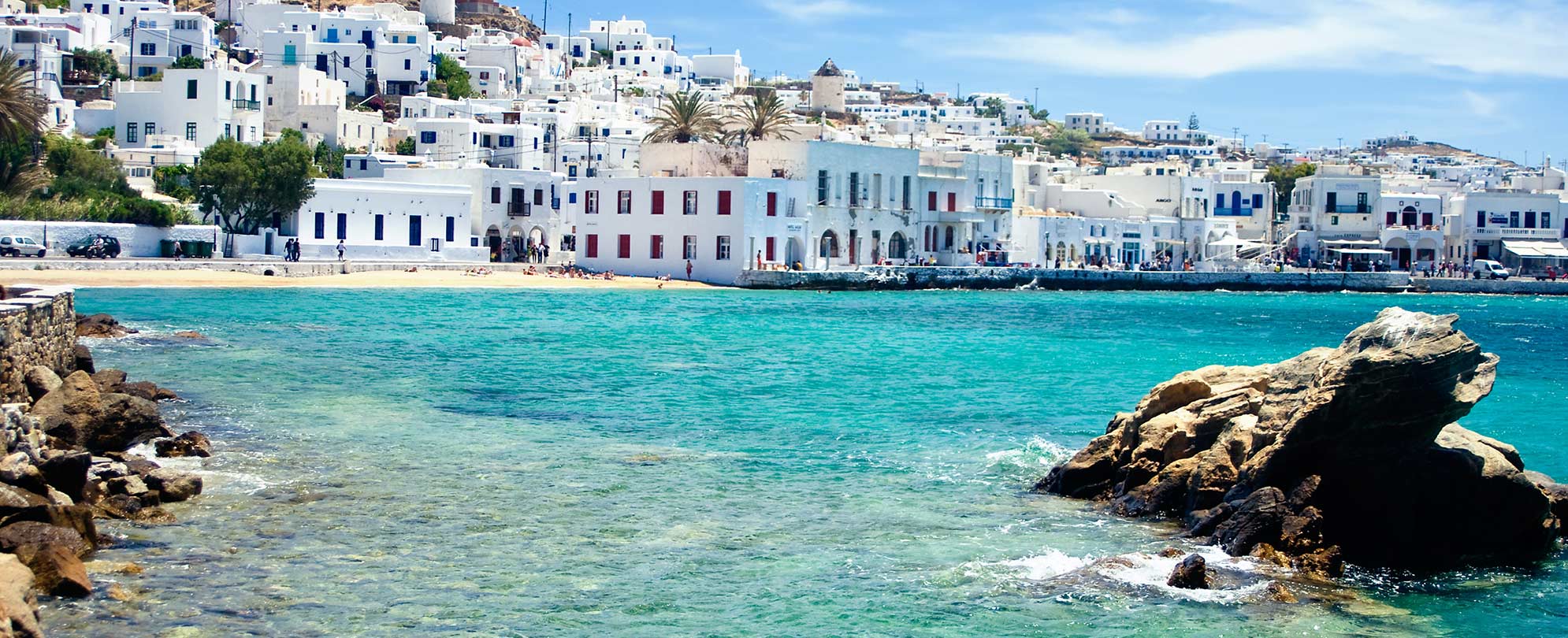 Sparkling water and white villas next to a rocky shoreline on a beach in Mykonos, Greece.