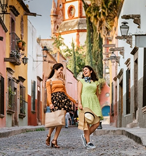 Two smiling woman holding bags walking on a cobblestone street with WorldMark by Wyndham Mexican cruise.