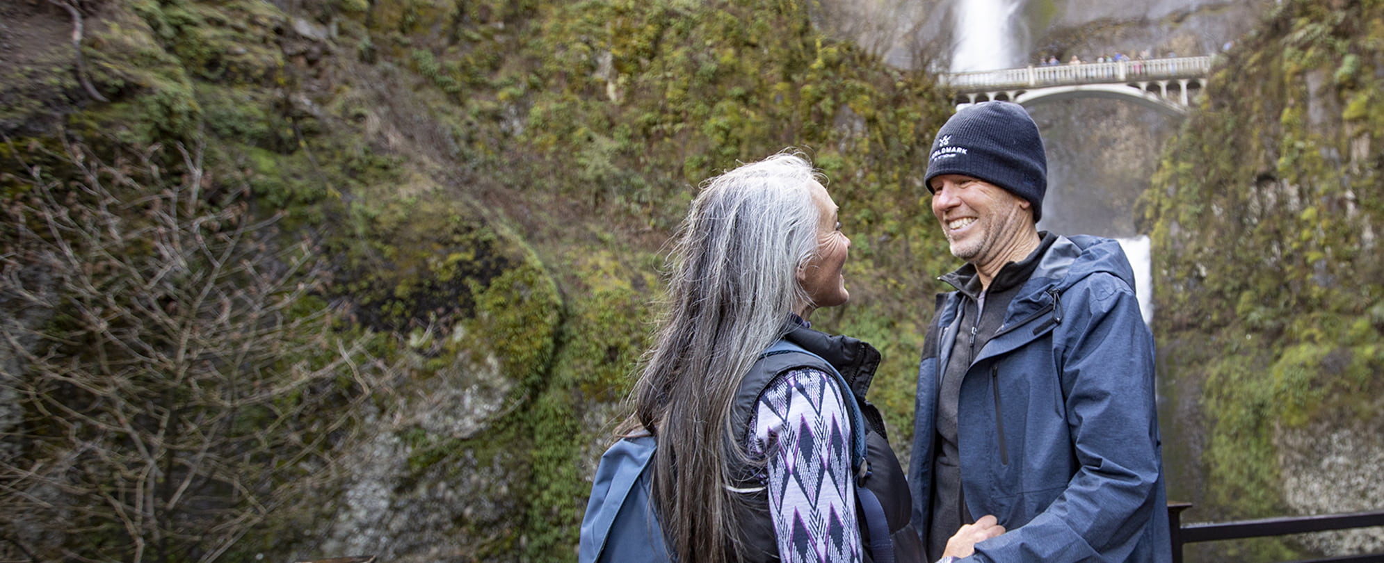 An older couple in WorldMark winter gear smile each other in front of a waterfall and bridge.