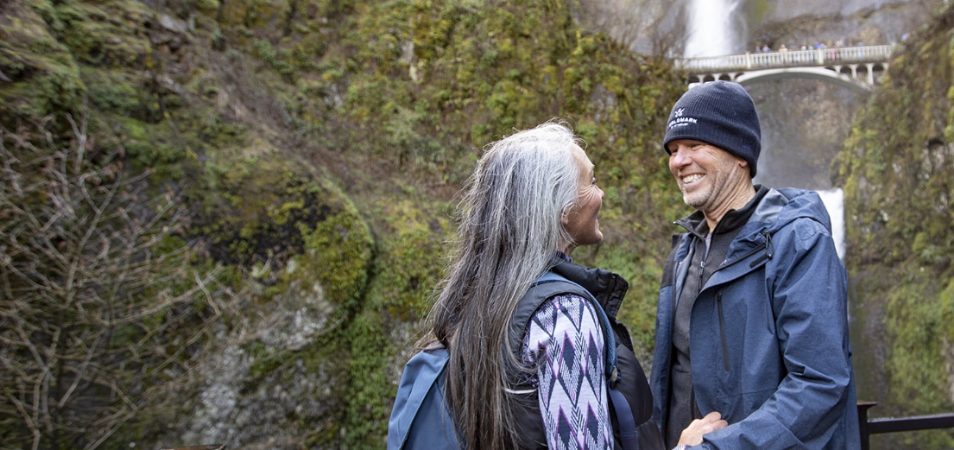 A man and woman walking side by side on a paved forest path, the woman is wearing a WorldMark by Wyndham backpack.