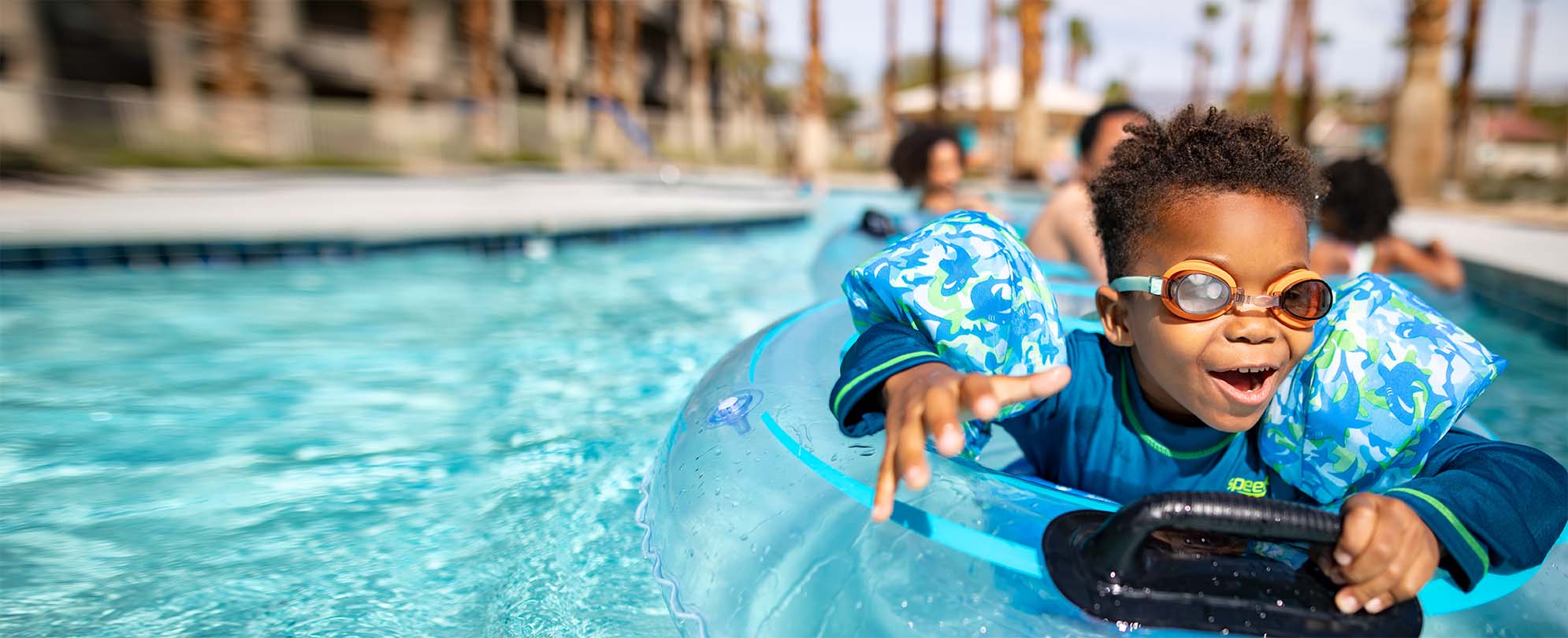 A smiling, young boy wearing swim goggles rides on a tube float at a WorldMark by Wyndham resort pool.