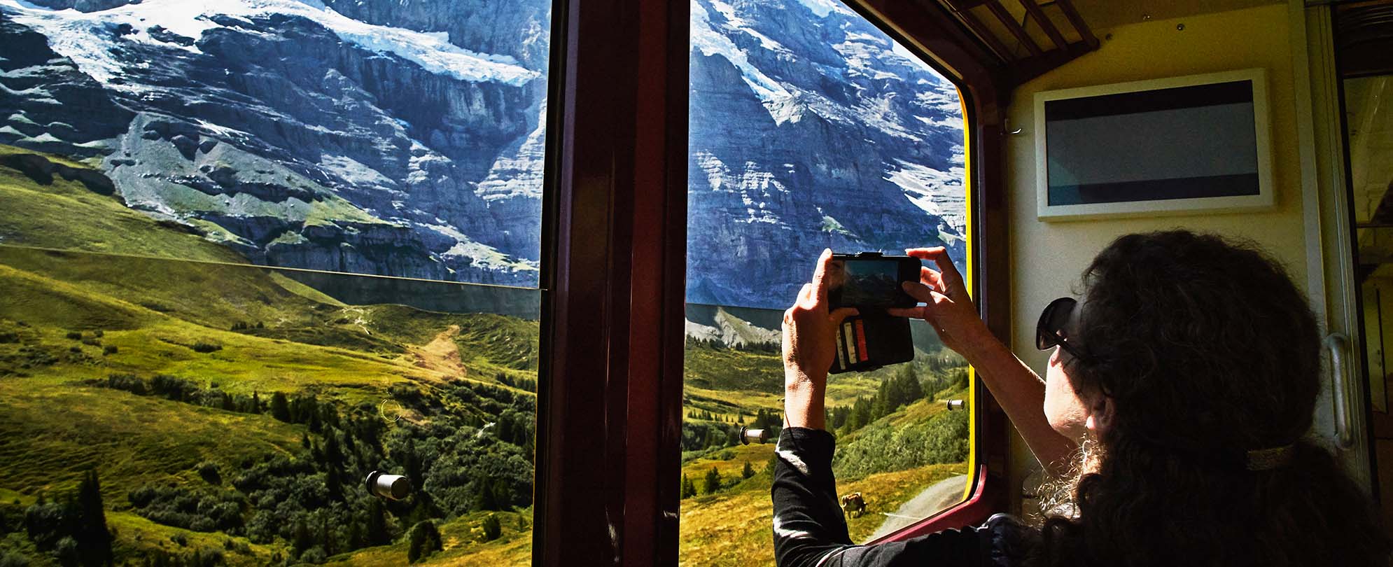 A woman, inside a train, taking a photo with her phone of the snow-covered mountains.