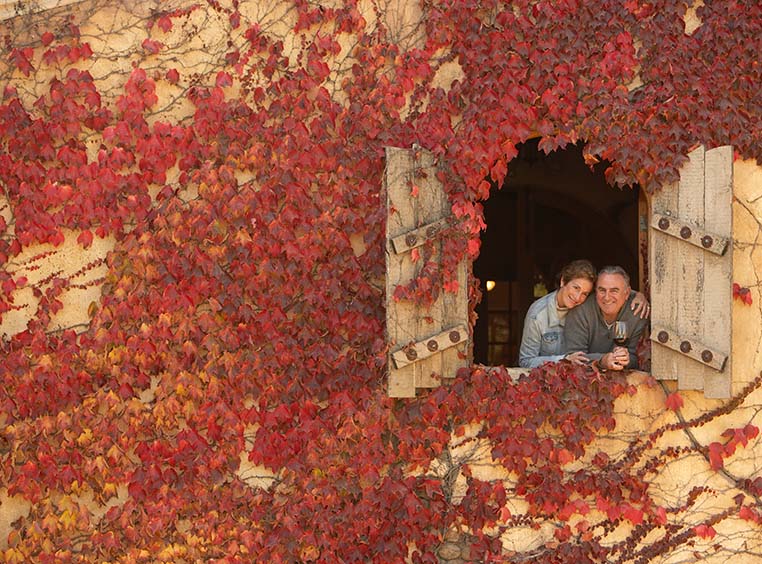 A couple, standing in the window of a European villa covered in red vines.