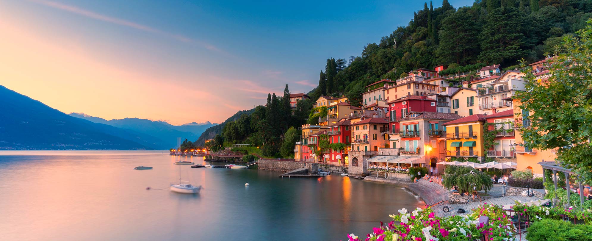 Colorful buildings on a mountainside overlooking Lake Como in Italy with bright skies and mountains in the distance.