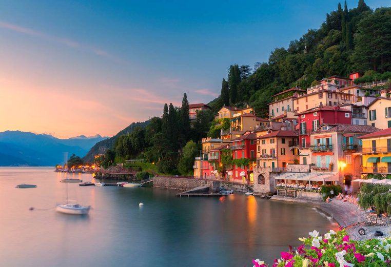 Colorful buildings on a mountainside overlooking Lake Como in Italy with bright skies and mountains in the distance.