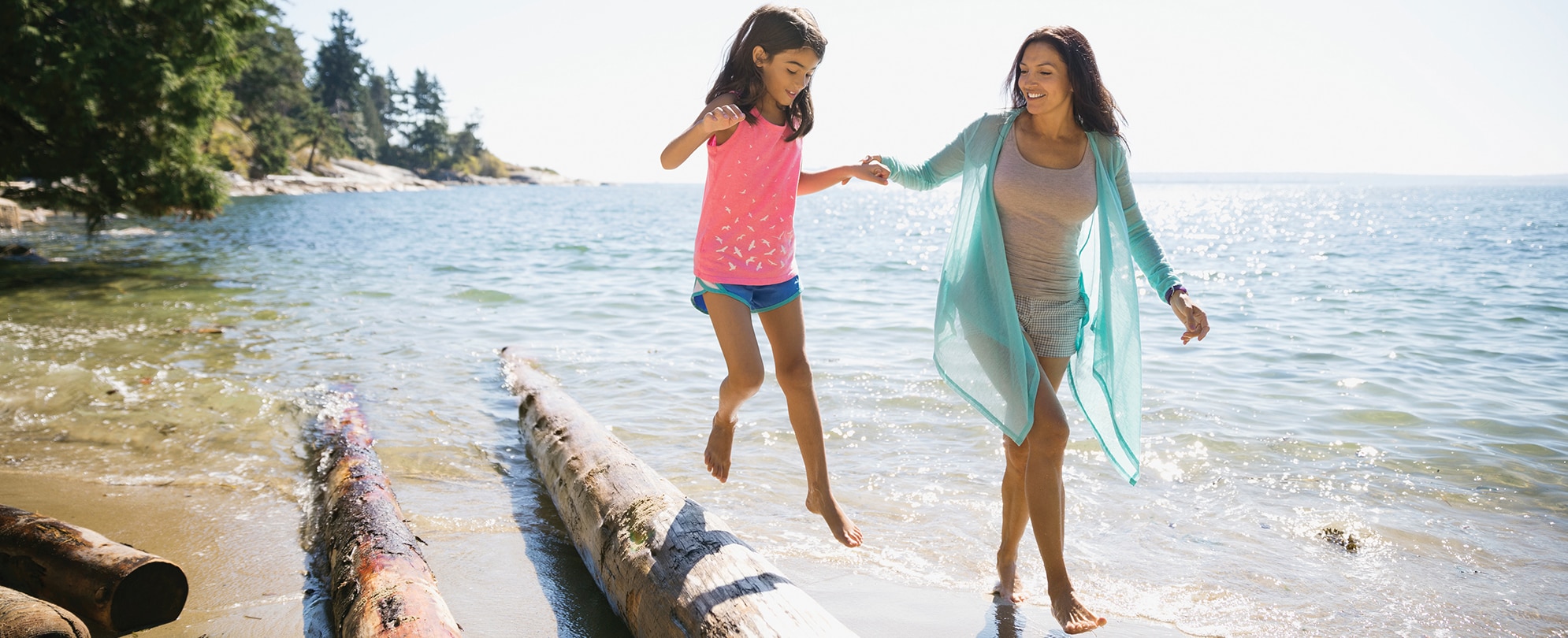 A mom holds her daughter’s hand as they walk on a beach, the daughter jumping off a fallen log by the water.