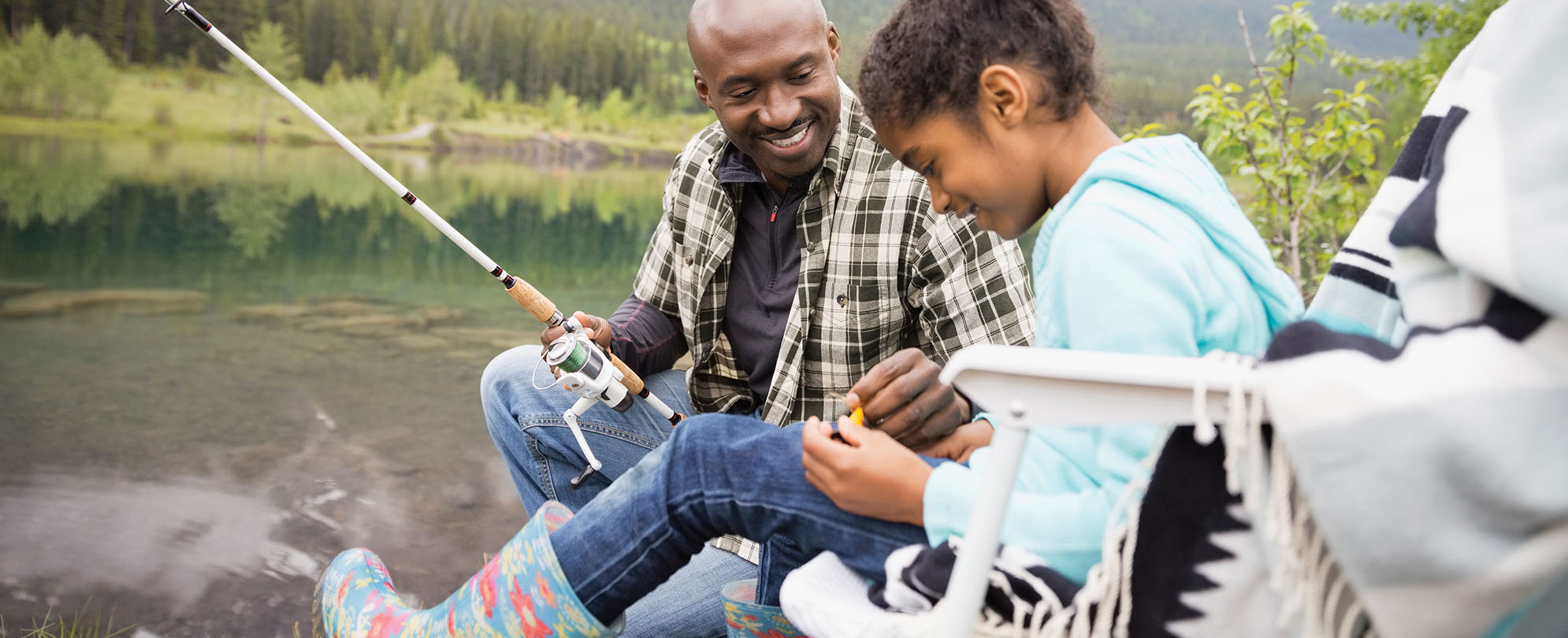 A dad and daughter fishing by the water, the smiling dad helping the daughter with a fishing hook.