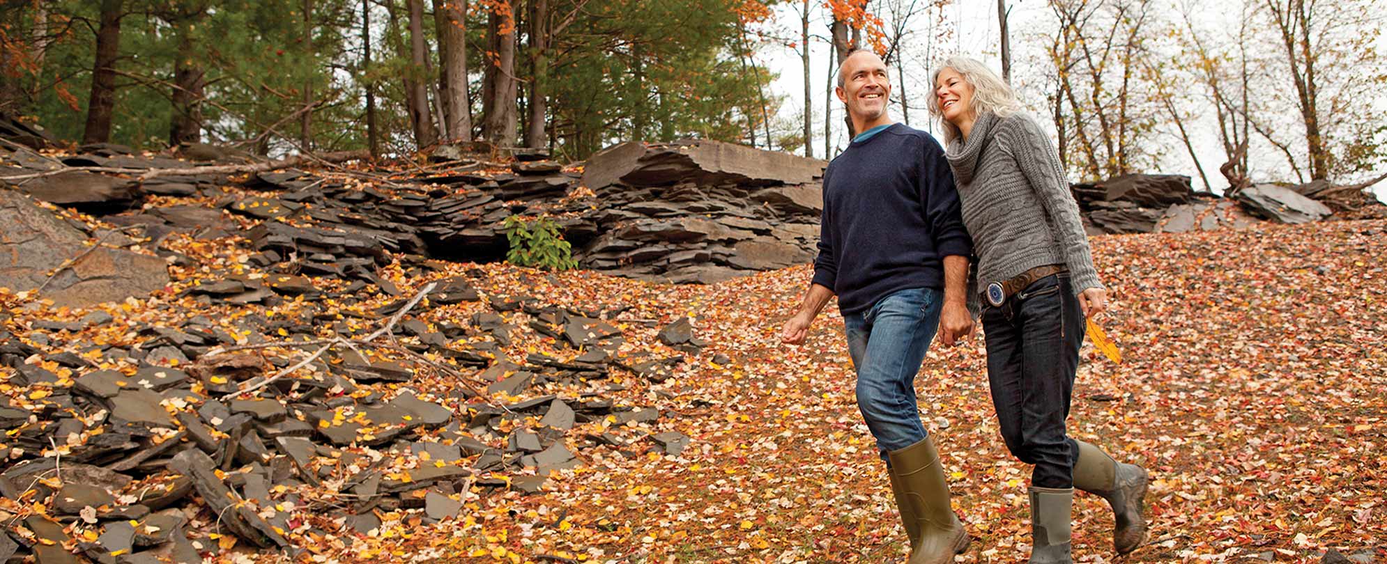An older man and woman holding hands walk down a trail covered in fall leaves.