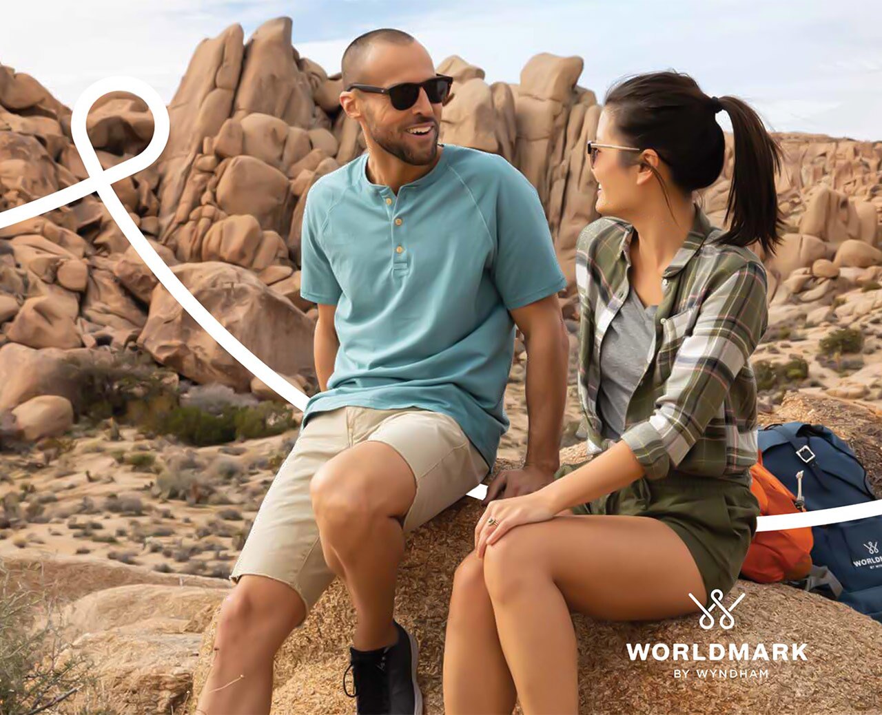 A man and woman wearing sunglasses smile at each other sitting on a boulder along a rocky hiking path.