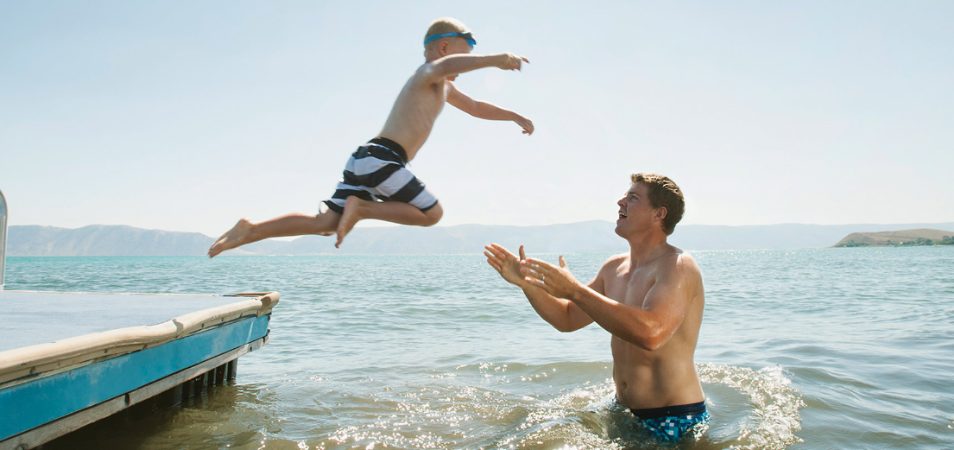 A young boy jumping off a dock as his dad stands in the ocean waiting to catch him.