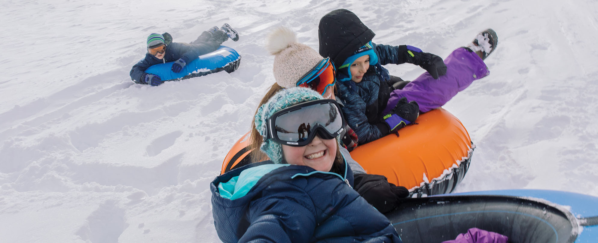 A smiling girl and her siblings wearing winter gear and goggles tubing in the snow.