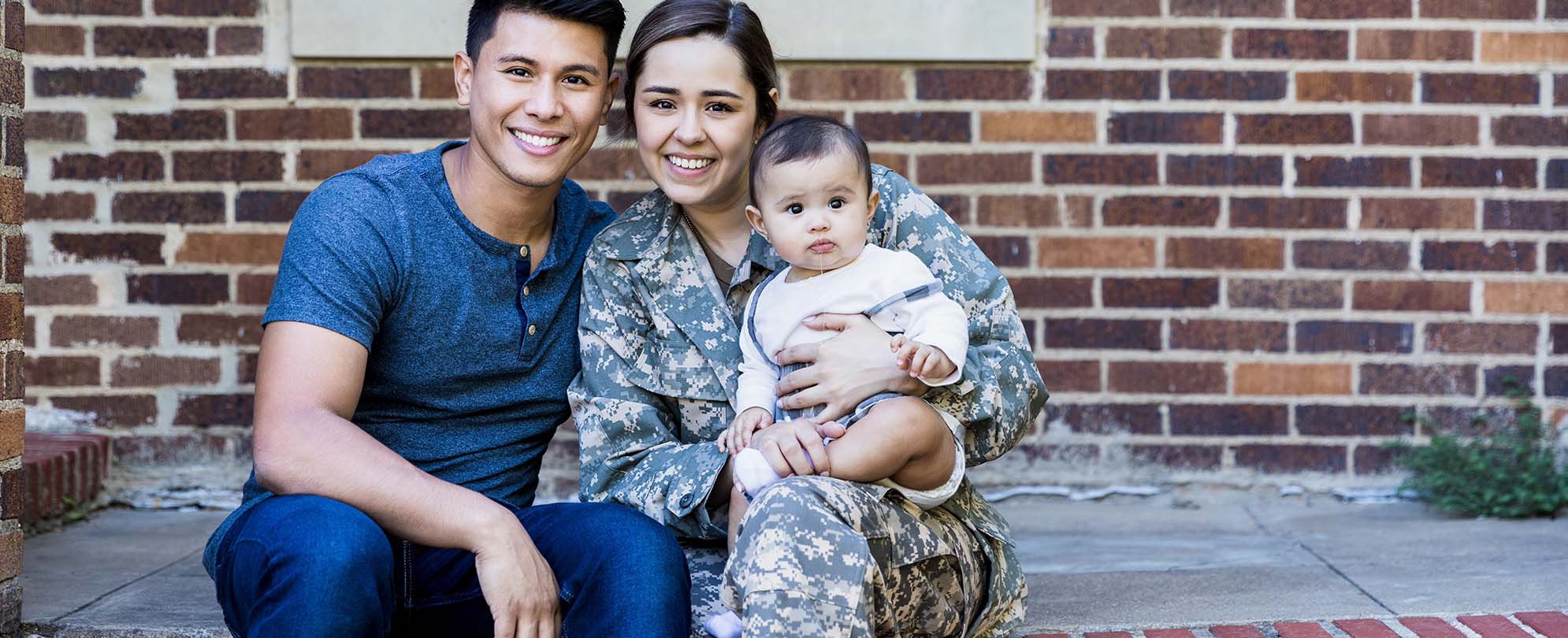 A woman in her military uniform, holding her baby while sitting next to her husband as they pose for a family photo. 