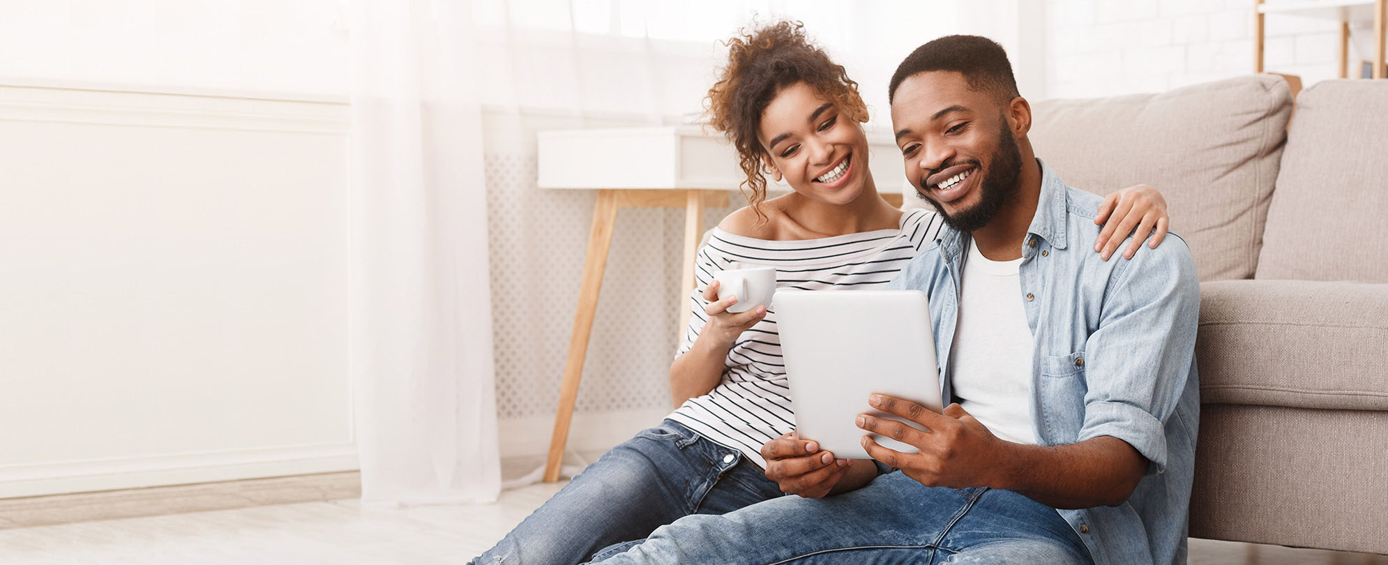 A couple sits comfortably on the floor in front of a couch, smiling down at a tablet.