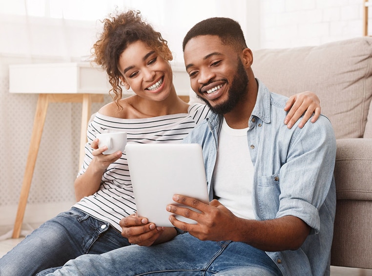 A couple sits comfortably on the floor in front of a couch, smiling down at a tablet.